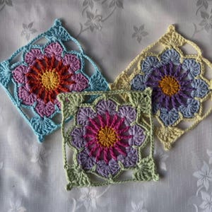 Peut inclure: Trois carrés au crochet faits à la main, aux motifs floraux. Chaque carré présente une fleur centrale et des pétales environnants dans des tons de bleu, rose, jaune, violet et vert. Ils sont disposés sur une surface blanche à motifs, mettant en valeur le travail de crochet.