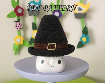 May include: A black crocheted pilgrim hat with a brown band and a yellow square buckle, resting on a white ceramic mug with a simple face design. The text PDF PATTERN is visible above the hat, which is set against a light background.