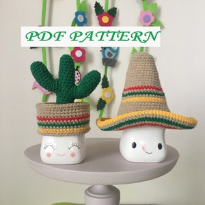 May include: Two crocheted cactus and sombrero ornaments. The cactus is green with a brown pot and red, yellow, and green stripes. The sombrero is brown with red, yellow, and green stripes. Both ornaments are sitting on a white mug with a happy face.