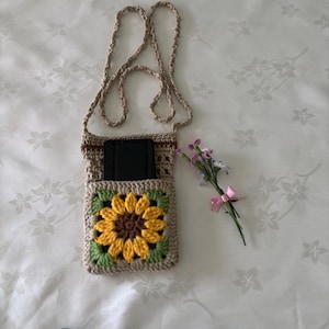 Op de afbeelding: Gehaakte crossbody telefoontas in beige met een zonnebloemontwerp. De tas heeft een vierkante zak met een gele en bruine zonnebloem, groene bladeren en een lange riem. Een klein boeket bloemen met een roze lint ligt naast de tas.