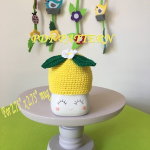 May include: Crochet lemon mug cozy pattern for a 2.75" x 2.75" mug. The cozy is yellow with green leaves and a white flower on top. The pattern is available as a PDF download.