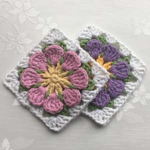 以下が含まれることがあります： 手作りのかぎ針編みの正方形が2つ。花柄のデザインです。各正方形は、ピンクまたは紫の花びら、黄色の中心、緑の葉を持つ中心の花を特徴とし、すべて白い背景に配置されています。正方形は、より大きなクラフトプロジェクトの一部である可能性があります。