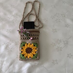 Op de afbeelding: Een beige gehaakte crossbody telefoontas met een zonnebloemontwerp. De tas heeft een gele en bruine zonnebloem in het midden, groene bladeren en een bruine band. Er zit een zwarte telefoon in de tas en er is een klein boeket bloemen bevestigd.