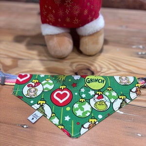 May include: Green Christmas-themed dog bandana featuring a Grinch and Cindy Lou Who design. The bandana has a red heart, green and white baubles, and the word "Grinch" in yellow lettering.