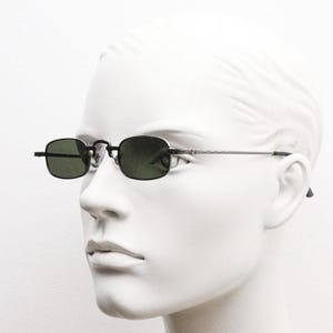May include: Rectangular sunglasses with dark green lenses and a black frame. The sunglasses have a thin, silver-colored arm that extends to the ear. The sunglasses are displayed on a white mannequin head.