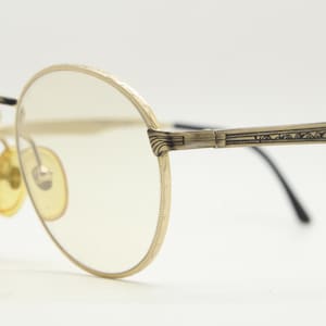 May include: Vintage gold-tone eyeglasses with oval lenses. The frames feature intricate detailing along the rims and temples, with a decorative pattern on the arms. The lenses are a light yellow tint, and the nose pads are also yellow.