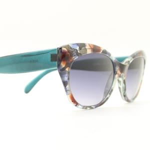 Puede incluir: Gafas de sol de ojo de gato con montura y patillas turquesas. Las monturas presentan un colorido estampado en marrón, azul y blanco. Las lentes tienen un tinte degradado, que va de un tono más oscuro en la parte superior a un tono más claro en la parte inferior.
