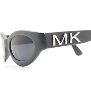 Puede incluir: Gafas de sol ovaladas negras con lentes oscuras. Las gafas tienen un acabado mate y presentan las letras "MK" en plata en las patillas. La montura tiene un diseño grueso y redondeado.