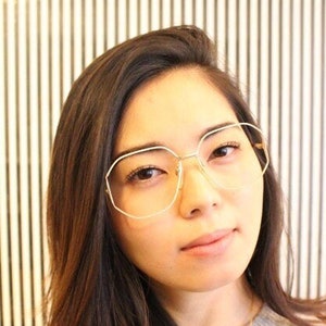 May include: A woman with long brown hair wearing clear, octagon-shaped glasses with a gold frame.