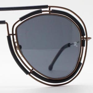 May include: Aviator-style sunglasses with a unique design. The sunglasses feature a black frame with a double-rimmed, circular design around the lenses. The lenses are a dark gray color. The bridge and arms are a bronze color.