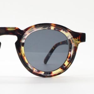 May include: Round sunglasses with a tortoiseshell pattern frame and dark gray lenses. The sunglasses have a keyhole bridge and black temples. The frame features a mix of brown, yellow, and black colors.