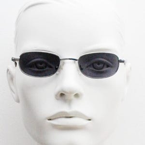 Puede incluir: Gafas de sol ovaladas con lentes oscuras y montura fina plateada. Las gafas se exhiben en una cabeza de maniquí blanca, resaltando el diseño. El estilo es minimalista y moderno, adecuado para el uso diario. El marco es de metal.