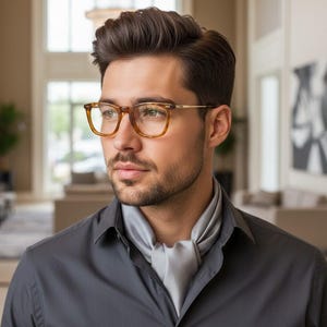 Puede incluir: Una persona con cabello oscuro y barba lleva puestas unas gafas rectangulares de color ámbar con patillas doradas finas y una camisa de cuello gris sólido con un pañuelo de seda gris a juego atado al cuello.
