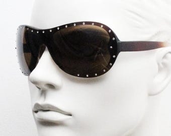 May include: Dark brown sunglasses with a curved frame and dark lenses. The frame is embellished with small, silver-colored studs. The temples transition from dark brown to a lighter brown color. The sunglasses are displayed on a white mannequin head.