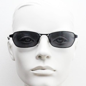 May include: Black rectangular sunglasses with dark lenses. The sunglasses have a thin, black frame and are displayed on a white mannequin head. The glasses are designed for eye protection from sunlight.