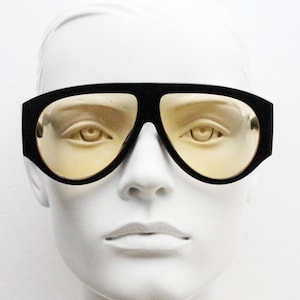 Może przedstawiać: Czarne okulary przeciwsłoneczne z żółtymi soczewkami. Okulary mają grubą, prostokątną oprawkę i są prezentowane na białej głowie manekina. Soczewki mają ciepły żółty kolor, a ogólny styl jest retro.