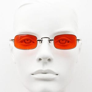 Może przedstawiać: Prostokątne okulary przeciwsłoneczne z cienkimi, srebrnymi metalowymi oprawkami i soczewkami w kolorze intensywnego pomarańczu. Okulary posiadają mostek łączący obie soczewki oraz smukłe zauszniki, przez które wyraźnie widać oczy użytkownika.