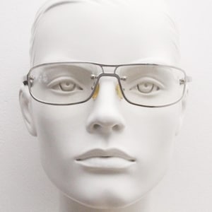 May include: Rectangular eyeglasses with a thin, dark metal frame and clear lenses. The glasses have a double bridge and are displayed on a white mannequin head. The nose pads are a light yellow color.