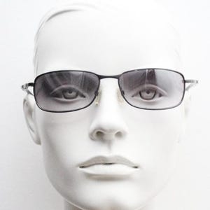 May include: Black rectangular sunglasses with a thin metal frame and gradient lenses. The sunglasses are displayed on a white mannequin head, showcasing the eyewear's design and fit. The lenses transition from dark at the top to lighter at the bottom.