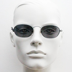 May include: A white mannequin head wearing oval-shaped sunglasses with dark grey lenses and a thin silver frame.