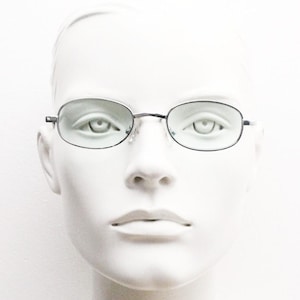 May include: Oval-shaped spectacles with a thin, dark metal frame. The lenses have a light green tint. The glasses are displayed on a white mannequin head, set against a plain white background. The overall aesthetic is minimalist and clean.