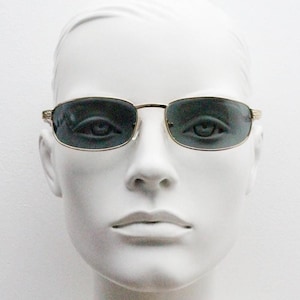 May include: A pair of gold-rimmed sunglasses with dark gray lenses. The sunglasses are on a white mannequin head.