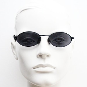 May include: Black oval sunglasses with a thin black frame. The sunglasses are displayed on a white mannequin head. The lenses are dark, obscuring the eyes. The overall style is minimalist and modern.
