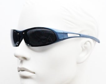 May include: Sport sunglasses with a dark blue frame and dark lenses. The sunglasses have a sleek, aerodynamic design with a curved shape and a cutout detail on the temples. The lenses are dark, providing protection from the sun.
