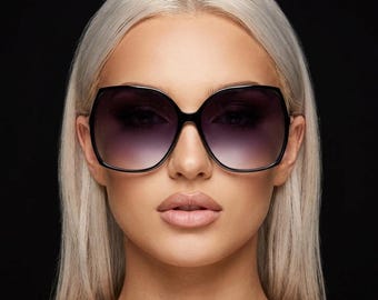 Puede incluir: Gafas de sol de gran tamaño con montura negra brillante y lentes degradadas que pasan de un tono oscuro en la parte superior a uno más claro en la inferior. La montura tiene una forma cuadrada redondeada con patillas finas y acabado liso.
