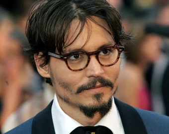 May include: A man with dark hair, wearing tortoiseshell eyeglasses and a dark suit with a black bow tie. He has a mustache and goatee. The background is blurred, suggesting an event or gathering.