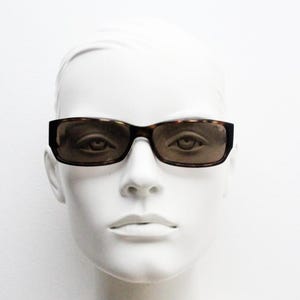 May include: Rectangular sunglasses with a tortoiseshell frame and tinted lenses. The sunglasses are displayed on a white mannequin head. The lenses are a dark brown color, and the frame has a mottled brown and black pattern.