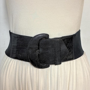 May include: A wide black belt with a large, decorative buckle. The belt has a textured, reptile-like pattern and an elastic back for a comfortable fit. The buckle is a prominent feature, adding a bold statement to the accessory.