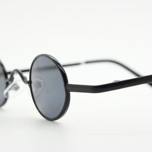 May include: Black round sunglasses with dark lenses. The frames are a matte black, and the lenses are a dark gray. The sunglasses have a classic, retro design, with a thin metal bridge and arms.