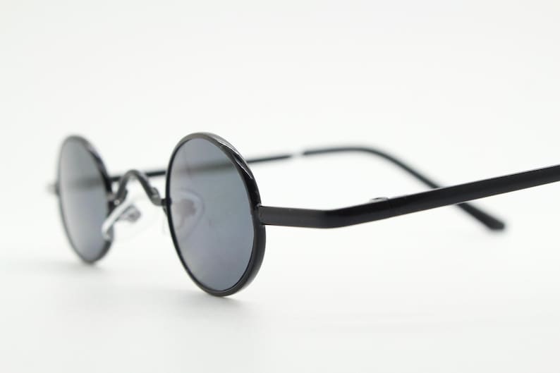 May include: Black round sunglasses with dark lenses. The frames are a matte black, and the lenses are a dark gray. The sunglasses have a classic, retro design, with a thin metal bridge and arms.