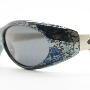 Op de afbeelding: Een paar blauwe en zilveren zonnebrillen met een bloemenpatroon op de monturen. De zonnebrillen hebben een zilveren metalen poot en een zilveren metalen logo aan de zijkant.