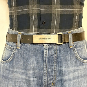 May include: Black leather belt with a silver-colored buckle. The buckle has the text "ARMAND BASI" engraved on it. The belt is worn with blue denim jeans and a blue and gray plaid shirt.