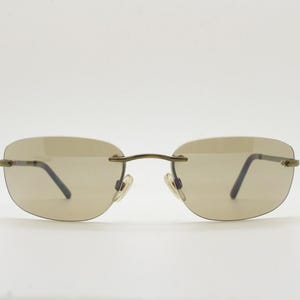 May include: Rimless sunglasses with a light brown tint. The glasses have a thin, gold-colored metal frame and dark temples. The lenses are oval-shaped and the nose pads are clear.