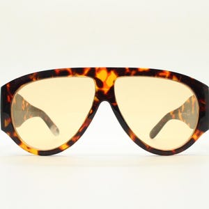 May include: Oversized sunglasses with a tortoise shell pattern frame and light yellow lenses. The sunglasses have a wide, rectangular shape with a prominent brow bar and thick temples. The lenses are a solid, uniform color.