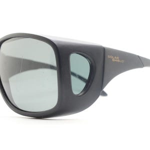 May include: Black sunglasses with wraparound frames and gray lenses. The sunglasses have a thick, protective design with the text "SOLAR SHIELD" printed on the temple.