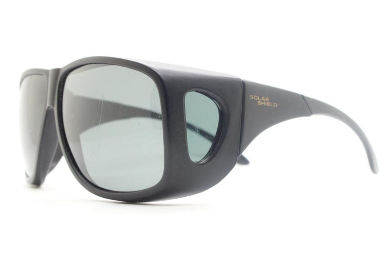 May include: Black sunglasses with wraparound frames and gray lenses. The sunglasses have a thick, protective design with the text "SOLAR SHIELD" printed on the temple.