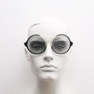 Könnte beinhalten: Ein weißer Mannequin-Kopf mit runden, schwarz umrandeten Sonnenbrillen mit grauen Gläsern.