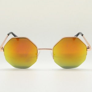 May include: Octagonal sunglasses with a rose gold frame and mirrored orange lenses. The sunglasses have a modern, geometric design with a sleek, minimalist aesthetic. The lenses transition from yellow to orange.