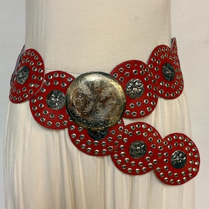 Cinturón vintage de los 80 para mujer, de cuero rojo con enormes círculos. Una pieza versátil y llamativa con tachuelas perforadas, discos de metal y una gran hebilla en forma de escudo. Nuevo sin usar.