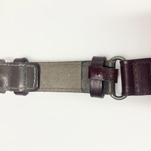 May include: A close-up of a brown leather belt with a dark metal buckle and a silver metal ring. The belt has a multi-toned design, with sections of gray, brown, and burgundy leather. White stitching is visible near the buckle.