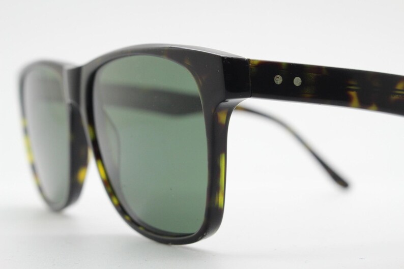 Puede incluir: Gafas de sol de carey negro y marr&oacute;n con lentes verdes. Las gafas tienen una forma rectangular cl&aacute;sica y un fino marco de metal.
