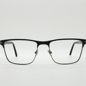 May include: Black rectangular eyeglasses with a thin metal frame and clear lenses. The glasses have a classic design with a bridge and nose pads. The temples are thin and black. The overall style is modern and minimalist.