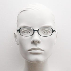May include: Black oval-shaped eyeglasses with a thin frame are displayed on a mannequin. The glasses have a subtle, minimalist design. The brand name, KOOKAI, is printed on the right lens.