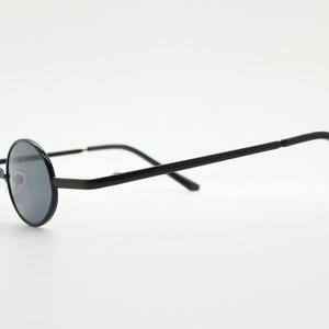 May include: Black round-frame sunglasses with dark lenses. The glasses have a thin, black metal frame and temples. The lenses are circular and the nose bridge is a silver color. The sunglasses are set against a white background.