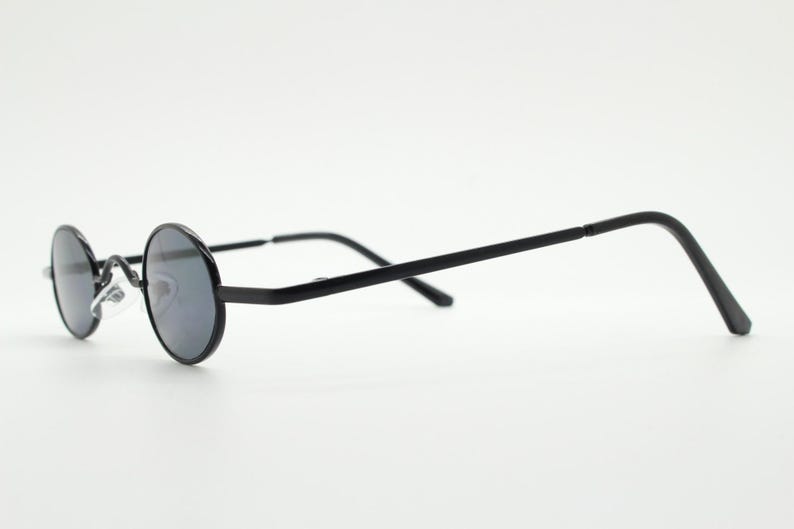 May include: Black round-frame sunglasses with dark lenses. The glasses have a thin, black metal frame and temples. The lenses are circular and the nose bridge is a silver color. The sunglasses are set against a white background.