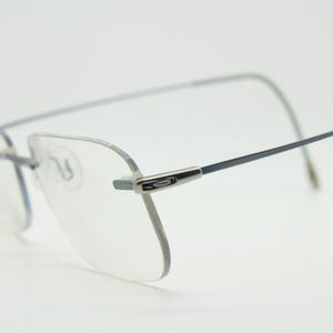 May include: Rimless spectacles with clear lenses and thin, silver-toned frames. The temples are a dark grey colour. The spectacles are designed for vision correction and are set against a white background.